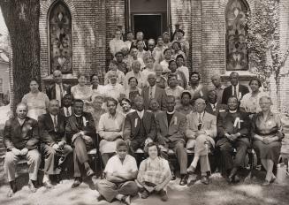 Black-and-white photograph of a large group of adults and children gathered in two rows outside…
