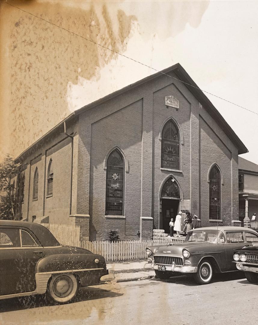 Black-and-white photograph of a one-storey brick church with a gable roof and tall, pointed-arc…