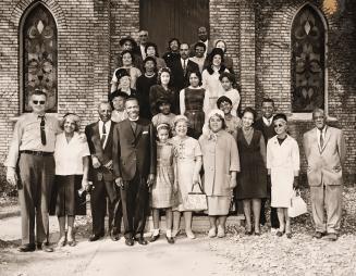 Black-and-white photograph showing 29 people posed on the steps of a brick building with arched…