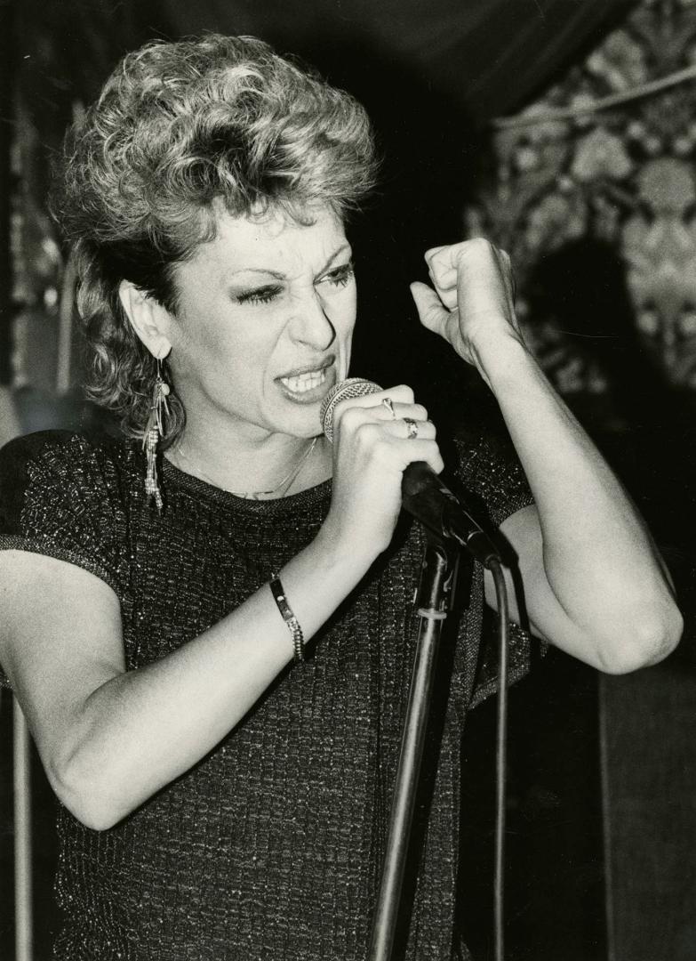 Woman in Sequin top and dangly earrings singing into hand held microphone with vigour. 