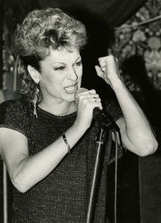 Woman in Sequin top and dangly earrings singing into hand held microphone with vigour. 