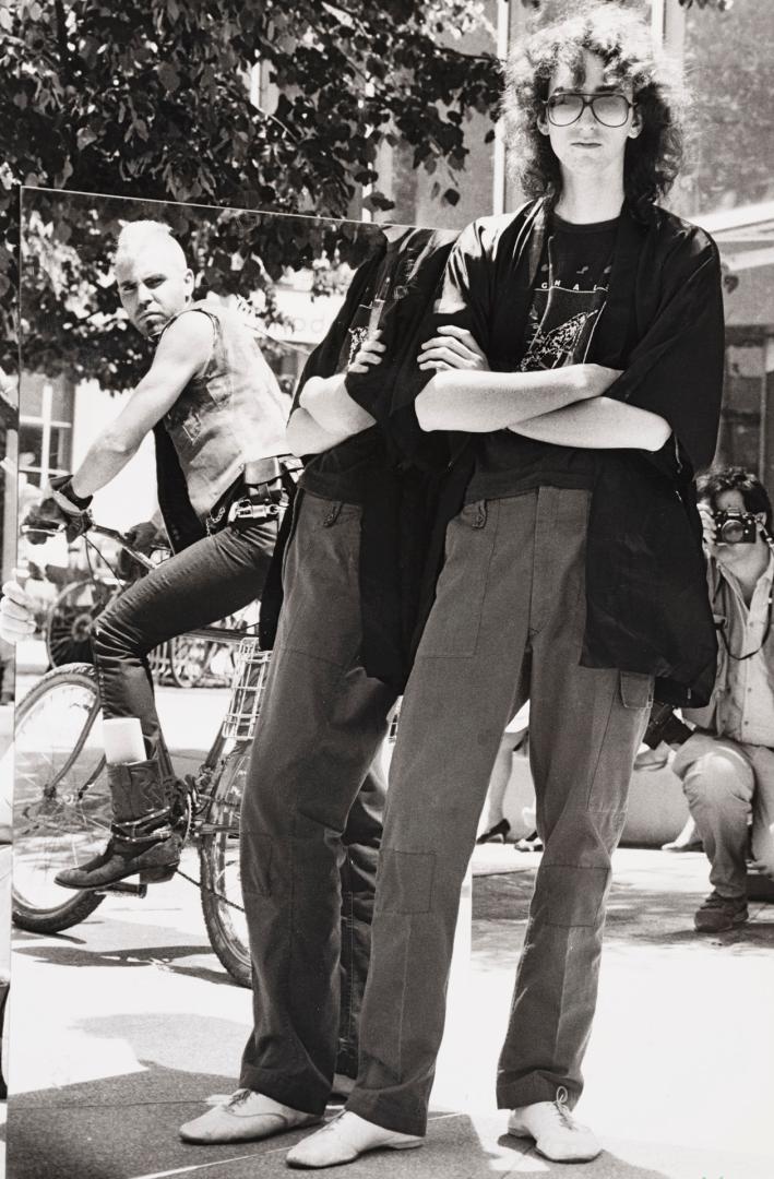 Outdoors in sunlight. Punk guy on bike is sneering at tall skinny young man with long curly hai…