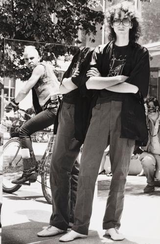 Outdoors in sunlight. Punk guy on bike is sneering at tall skinny young man with long curly hai…