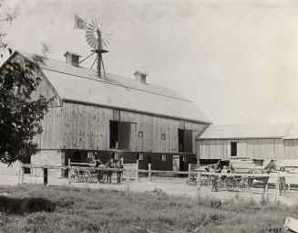 Black and white photograph of a barn. In front of the barn are two horse drawn buggies with peo…
