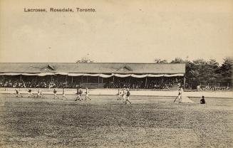 Grey-toned postcard depicting a lacrosse field, players, and audience in the viewing stands. Th ...
