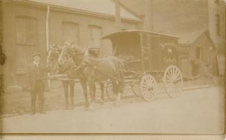 Black and white photograph of a man standing in front of a horse-drawn wagon.