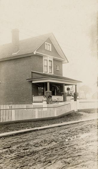 Black and white photograph of a two story house with a picket fence around it. Two lades are po ...